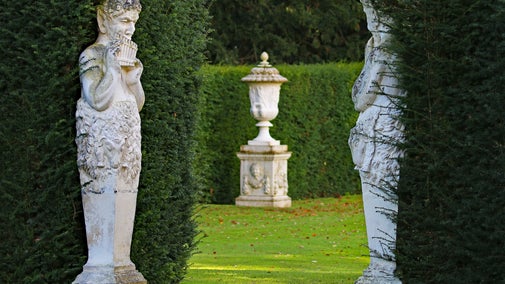 A satyr statue is playing the panpipes by a hedge, the entrance to the Rose Garden. An urn can be seen in the gap between the hedge.